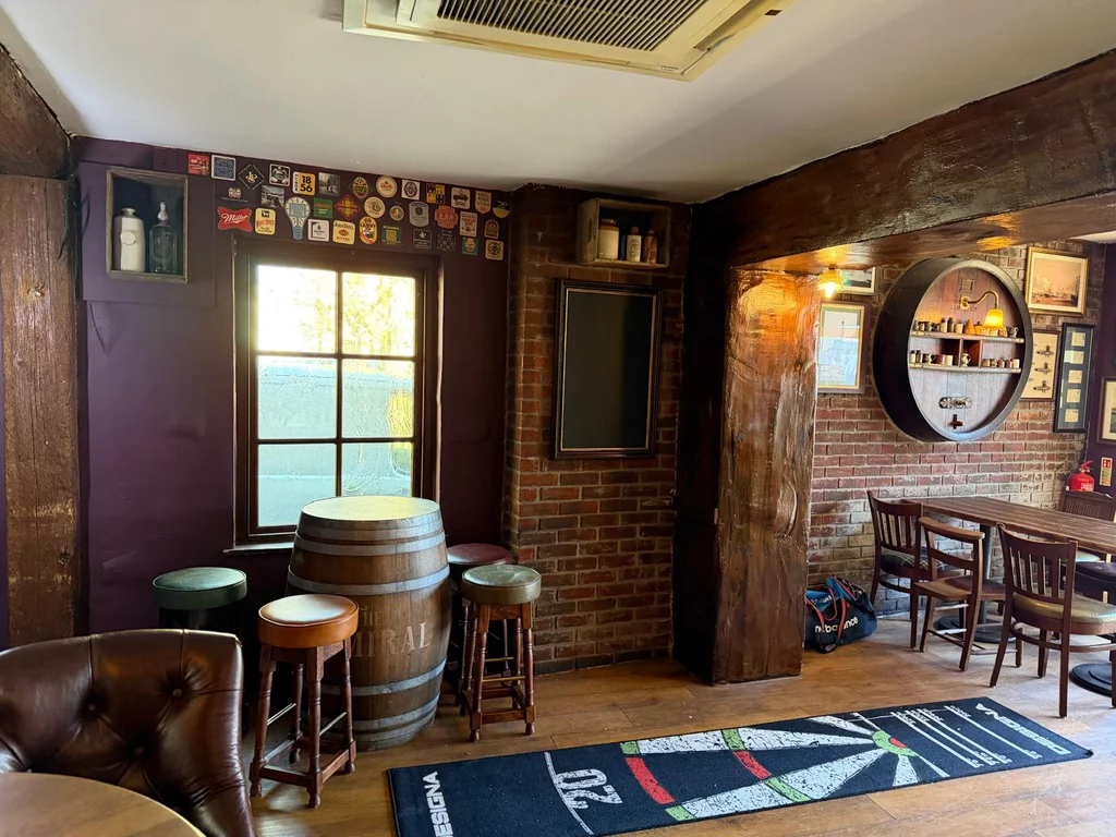 The image shows a cozy bar with rustic charm, featuring a wooden table, two bar stools, a wooden barrel, a round mirror, a television, and a wooden floor. The brick wall has a photo frame, a vent, and a window with a view of the ocean. The wooden floor and bar stools create a comfortable atmosphere, while the mirror and television add modern touches. The scene is well-organised and inviting, perfect for socializing and enjoying a drink.