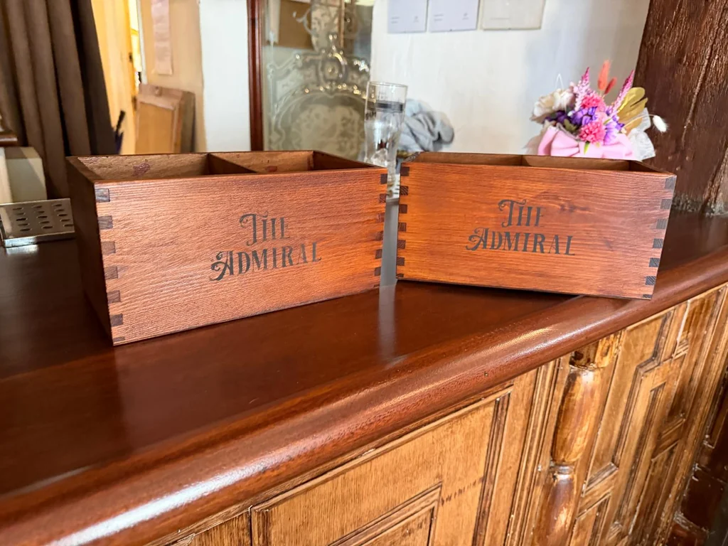 The image features a wooden counter with two wooden crates labeled "The House" and "The Garden" on it, placed in front of a mirror and a vase of pink and white flowers. The scene is set against a wooden cabinet with a mirror and a window, creating a harmonious and inviting atmosphere.