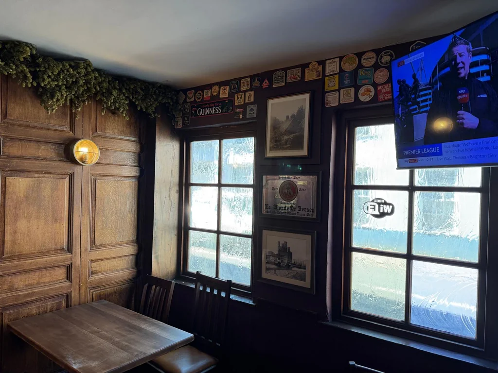 The image shows a cozy, rustic bar with a wooden table, chairs, and a green wreath. The walls are adorned with various posters and pictures, including a beer advertisement. A television is mounted on the wall, and a yellow light hangs above the table. The room is well-lit, creating a warm and inviting atmosphere.