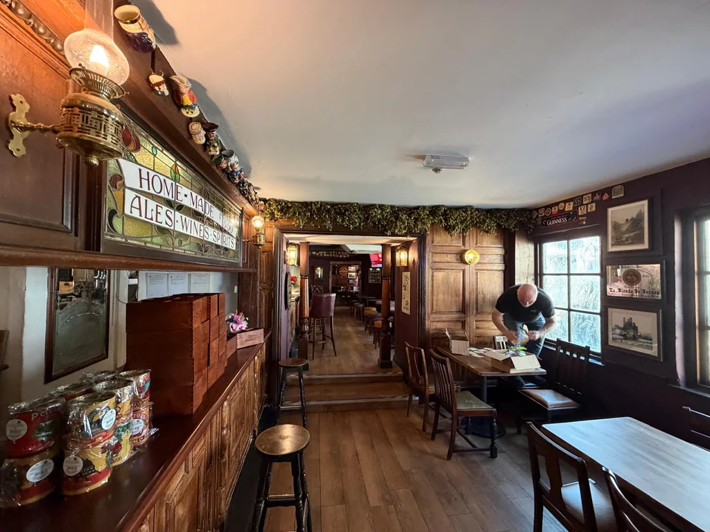 The image shows a cozy, rustic bar with wooden walls and a wooden counter. The counter has a "Brewery" sign and a "Brewery's Bar" sign. The bar is filled with various items, including bottles, cans, and jars. The floor is wooden, and the walls are decorated with greenery and pictures. A man is standing near the bar, and a stool is placed in front of it. The bar is well-lit, and the overall atmosphere is warm and inviting.