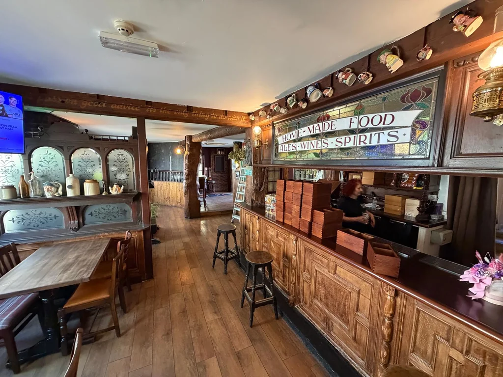 The image shows a cozy, rustic bar with wooden floors and walls, featuring a wooden counter, stools, and a bar with a large sign. The bar is illuminated by hanging lamps and has a blue ceiling fan. The wooden elements create a warm and inviting atmosphere, and the bar is well-stocked with various items. The scene is a blend of rustic charm and modern convenience.