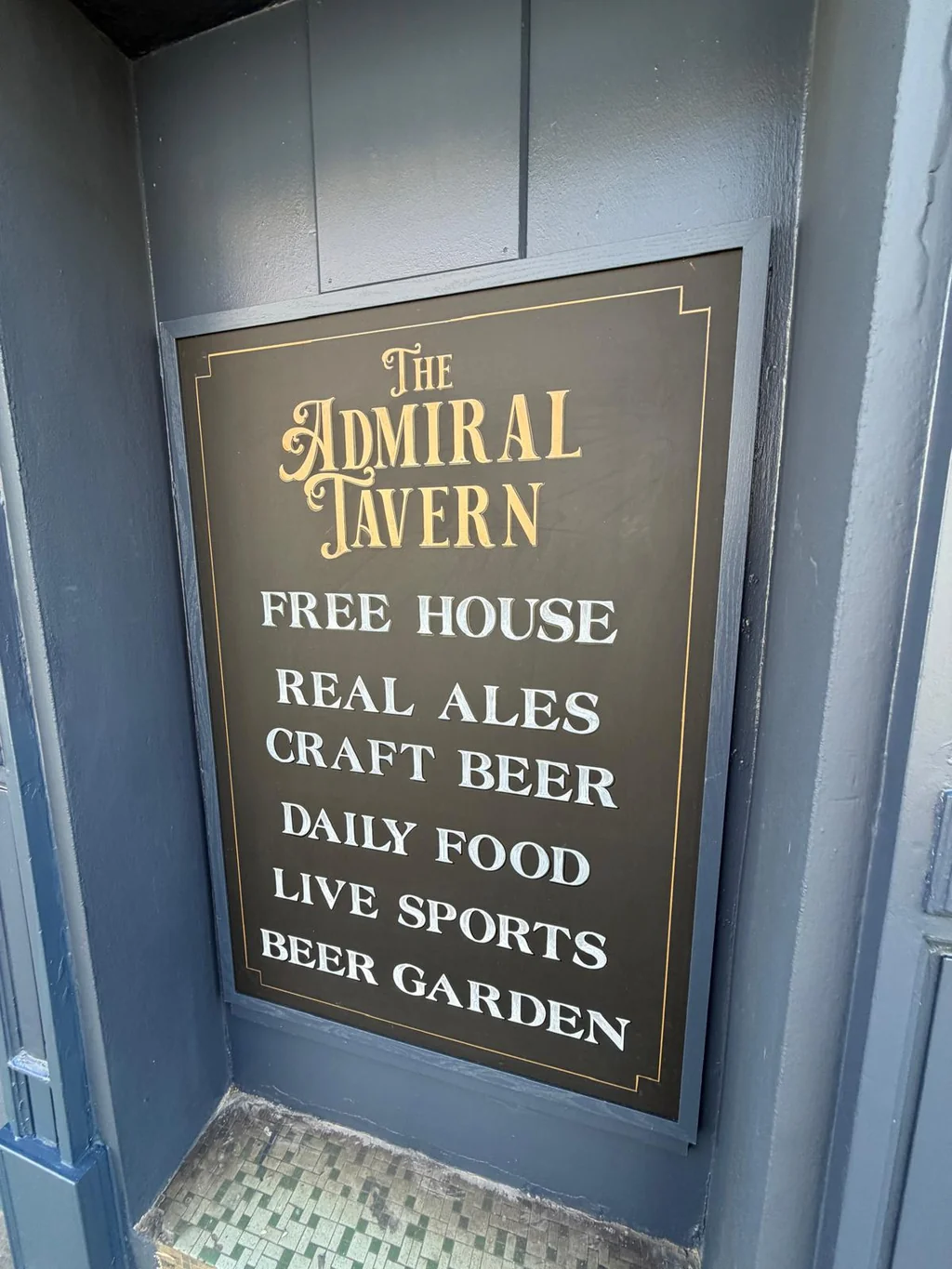 The image features a black sign with gold text on a blue wall, displaying the "The Mill Inn" name and a list of beers, including "Black & Gold," "Red & Gold," "Red & Black," "Black & Black," and "Black & Gold." The sign is mounted on a blue door, and the image is taken from a low angle, emphasizing the sign's size and importance.