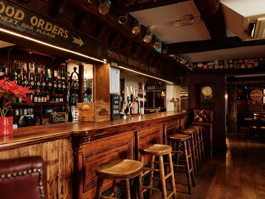 The image shows a cozy, rustic bar with a wooden counter, bar stools, and a red-potted plant. The counter has a black sign with a yellow arrow, and a wooden clock hangs above. The bar is filled with various liquor bottles and a red vase with red flowers. The floor is made of dark wood, and the ceiling has a wooden beam. The atmosphere is warm and inviting, with a sense of history and comfort.
