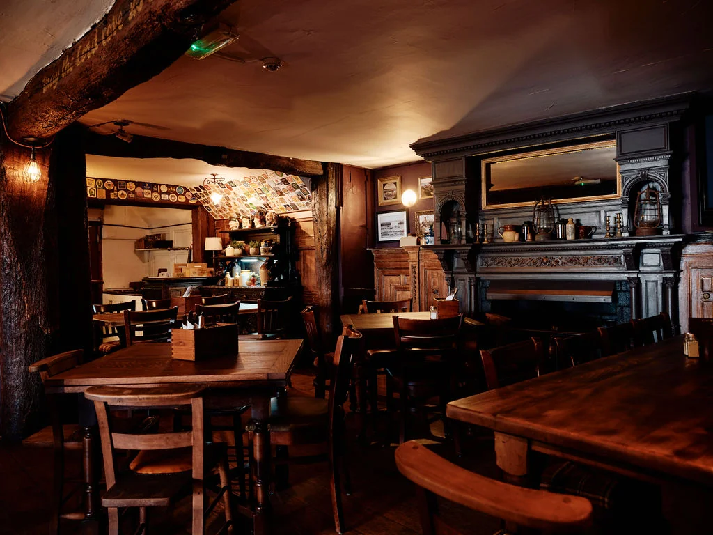 The image shows a cozy, rustic bar with dark wood furniture, a wooden bar, and a fireplace. The bar has a wooden counter and a mirror above it. The walls are adorned with a mirror, a clock, and a picture. The ceiling has a wooden beam and a hanging light fixture. The floor is dark wood, and the atmosphere is warm and inviting.