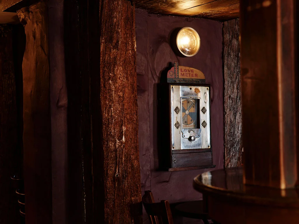 In a dimly lit room, a vintage arcade machine with a gold-colored dial and a black screen displays a game with a yellow circle and a red circle. A wooden table and chair are to the right, and a wooden wall with a large tree trunk is on the left. The scene is bathed in warm, soft light, creating a nostalgic atmosphere.