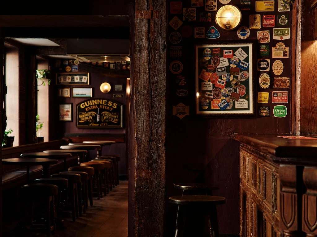 The image shows a cozy, rustic bar with dark wood paneling, a wooden counter, and stools. The wall features beer advertisements, a "Brews" sign, and a hanging lamp. The floor is wooden, and the perspective is from the entrance, inviting patrons to enjoy the atmosphere.
