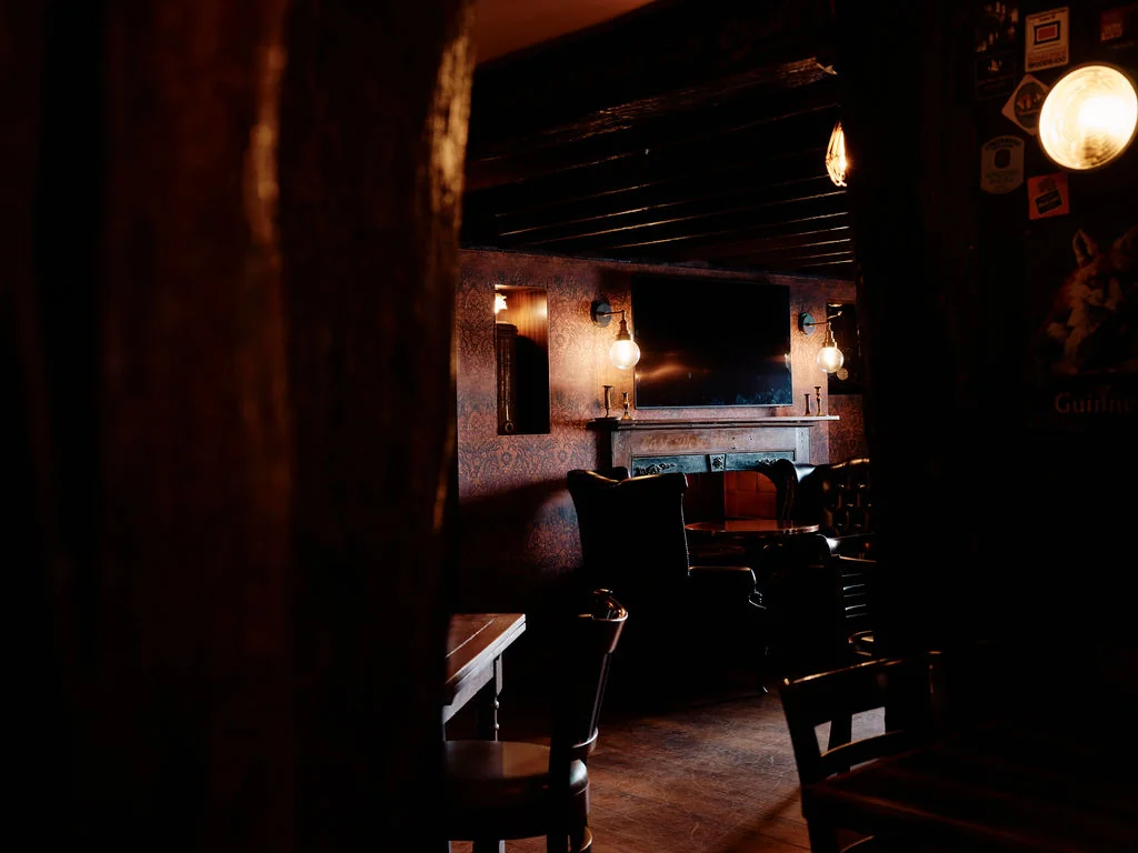 In a dimly lit bar, a wooden table with two chairs and a wooden bar is illuminated by three hanging lamps. A large mirror hangs above the table, and a wooden tree trunk is on the left wall. The floor is made of wood, and the atmosphere is cozy and inviting.