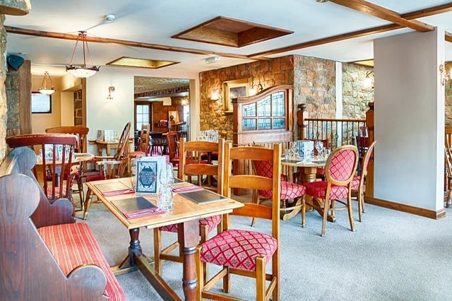 The image features a beautifully set dining table with a large, dark brown steak as the centerpiece, surrounded by a variety of food items. The steak is garnished with a red sauce and is accompanied by a plate of roasted potatoes, a plate of broccoli, and a plate of carrots. A glass of wine and a silver salt shaker are also present, along with a white napkin and a silver fork. The scene is set against a wooden table, creating a warm and inviting atmosphere.