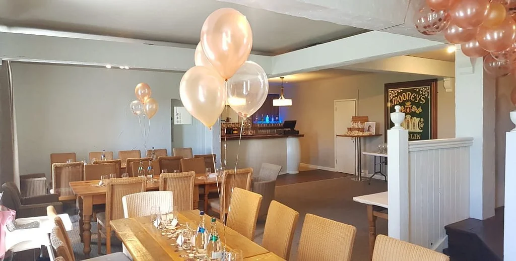 The image shows a spacious, modern restaurant with a warm, inviting atmosphere. The room is decorated with pink and white balloons, creating a festive mood. The wooden tables and chairs are set for a celebration, with silverware and napkins. A bar area with a blue sign and a red sign is in the background, and a white wall with a painting adds artistic flair. The room is well-lit, with hanging lights and a ceiling light. The arrangement of objects and decorations suggests a celebration of good times and good company.