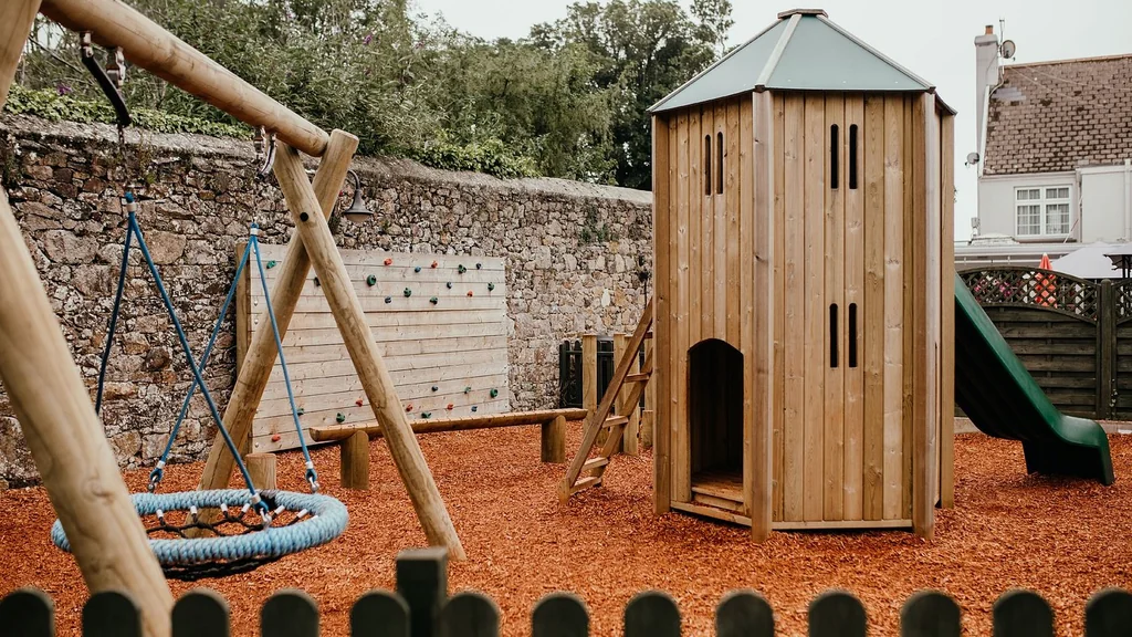 A wooden swing set with a blue swing and a green slide is in the foreground, surrounded by a wooden wall and a stone wall. The ground is covered in red mulch, and a green playground is in the background. The scene is peaceful and inviting, with a sense of safety and playfulness.