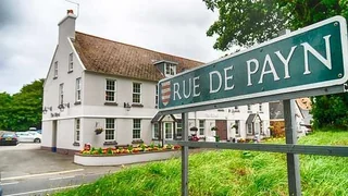 The image features a large, white two-story building with a red roof, surrounded by lush greenery and a clear blue sky. The building has multiple windows, a chimney, and a door. In front of the building, there are two parked cars and a green street sign with white lettering. The scene is set on a street with a white line and a grassy area.