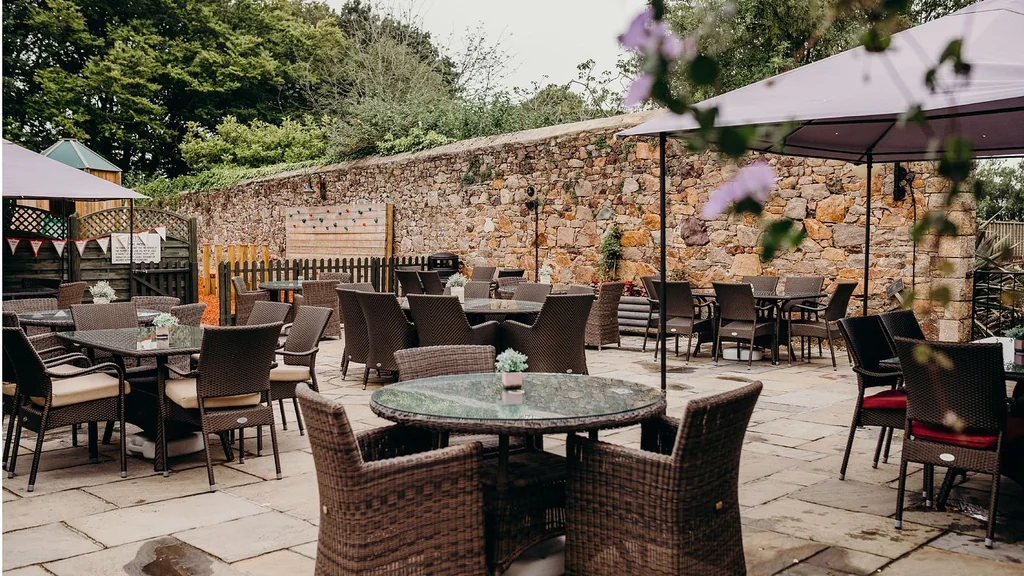 The image shows a serene outdoor patio area with six wicker chairs and tables, each accompanied by a green glass table. The tables are set under purple umbrellas, and the patio is surrounded by a stone wall and a wooden fence. The scene is bathed in soft, diffused light, creating a tranquil atmosphere.