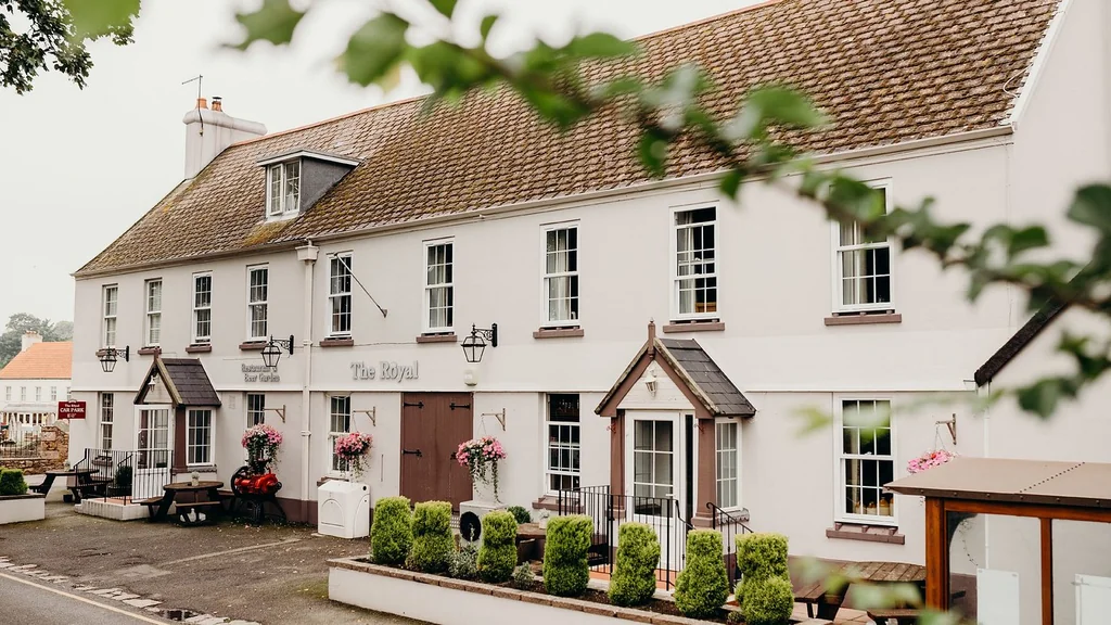 The image features a white two-story hotel with a red tiled roof, surrounded by lush greenery. The hotel has a "The Hotel" sign and a "Restrooms" sign, indicating it's a place for rest and relaxation. The street in front of the hotel is lined with trees and bushes, creating a serene atmosphere. The perspective is from the street, looking up at the hotel, emphasizing its grandeur and elegance.