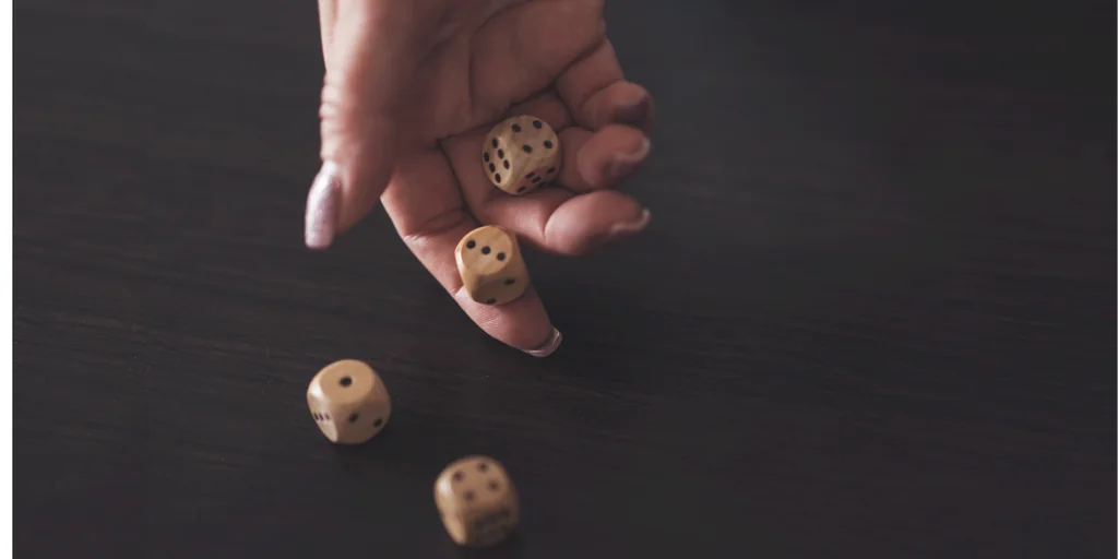 A hand with a light-colored nail polish is holding a pile of three light-colored dice on a dark wooden surface, with one dice slightly above the others. The background is blurred, emphasizing the hand and dice. The image captures a moment of anticipation, with the hand and dice as the main focus.