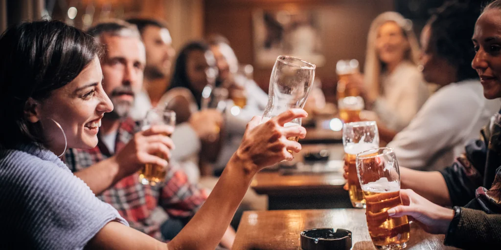 A lively bar scene features a group of people enjoying beer at a wooden table, with one person holding a large glass filled with beer. The background is filled with other patrons, and the atmosphere is warm and convivial.