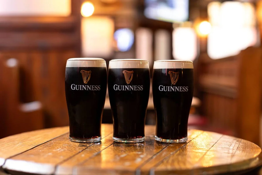 Three dark-colored Guinness glasses filled with dark beer are arranged in a triangle on a wooden table in a cozy pub or bar setting. The background is blurred, emphasizing the glasses and table. The image is taken from a low angle, giving a unique perspective. The image code "sa_15007" may refer to a specific landmark or location within the pub.