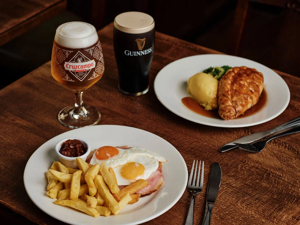 A cozy meal setting on a wooden table features two white plates with a sunny-side-up egg, golden fries, and a golden-brown ham and cheese sandwich. A dark-colored Guinness beer is to the right, and a dark-colored red sauce is on the left. The arrangement creates a harmonious and inviting atmosphere.