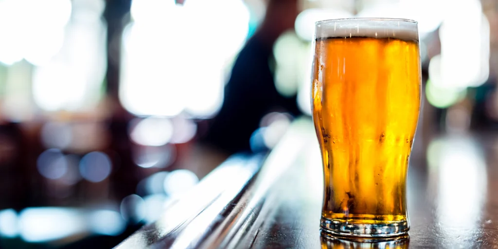 A tall, golden beer glass filled with a frothy yellow beer sits on a wooden bar, with a blurred background of other patrons and a blurred person. The glass is the main focus, with the background elements providing context.