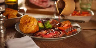 The image features a beautifully set dining table with a large, dark brown steak as the centerpiece, surrounded by a variety of food items. The steak is garnished with a red sauce and is accompanied by a plate of roasted potatoes, a plate of broccoli, and a plate of carrots. A glass of wine and a silver salt shaker are also present, along with a white napkin and a silver fork. The scene is set against a wooden table, creating a warm and inviting atmosphere.