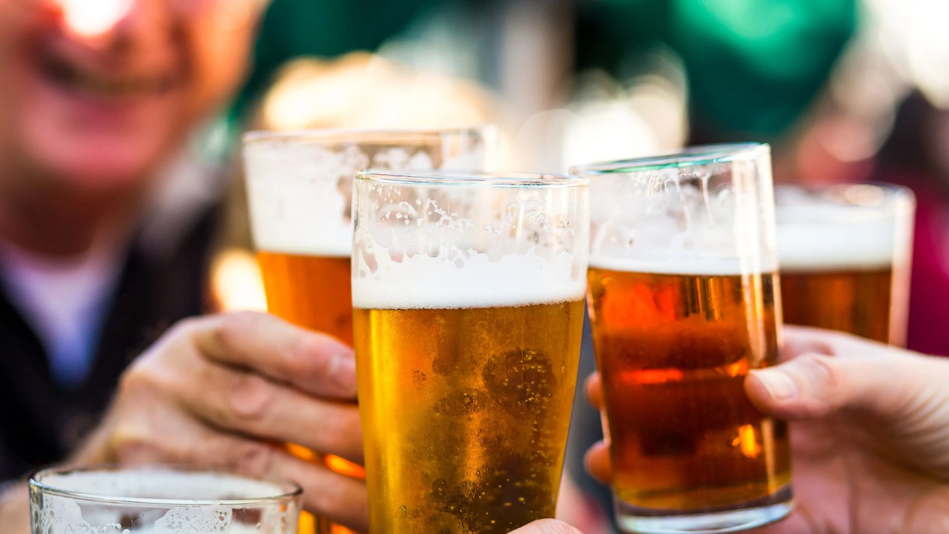 Four people are gathered at a table, each holding a glass filled with a golden-brown beer. The glasses are filled to the brim, with the beer frothy and golden. The background is blurred, suggesting a lively social setting. The image captures a moment of camaraderie and enjoyment.