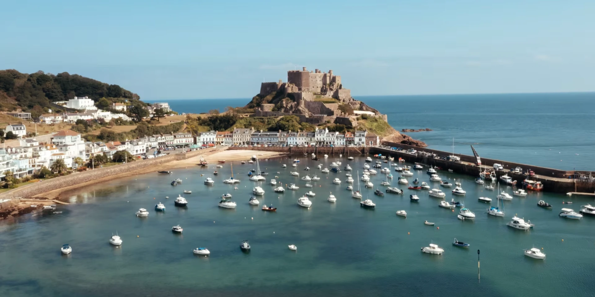 The image features a serene harbor scene with a large number of boats docked in the water, surrounded by white buildings. A castle-like structure stands on a hill, overlooking the harbor and buildings. The sky is a clear blue with a few clouds, and the overall scene is harmonious and tranquil.