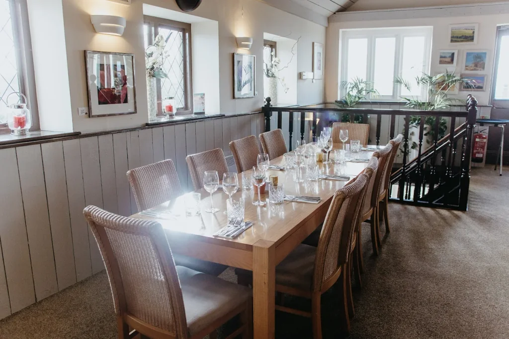 The image shows a cosy restaurant with a long wooden table set for six, surrounded by white chairs with blue cushions. The table is adorned with white plates, silverware, and white napkins. The walls are decorated with framed pictures and a black railing. The floor is carpeted, and the room is well-lit by natural light. The arrangement of objects and the careful placement of furniture create a harmonious and inviting atmosphere.