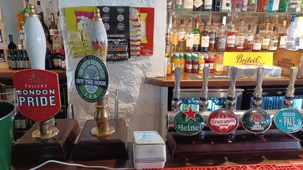 The image shows a well-stocked bar with six beer taps on wooden counters, each with a green label and a red label. A yellow sign with "Brewin's" and "Brewin's" is on the wall behind the taps. The counter is white, and the background features a white wall with a poster of beer bottles. The arrangement of the taps and the poster create a visually appealing scene.
