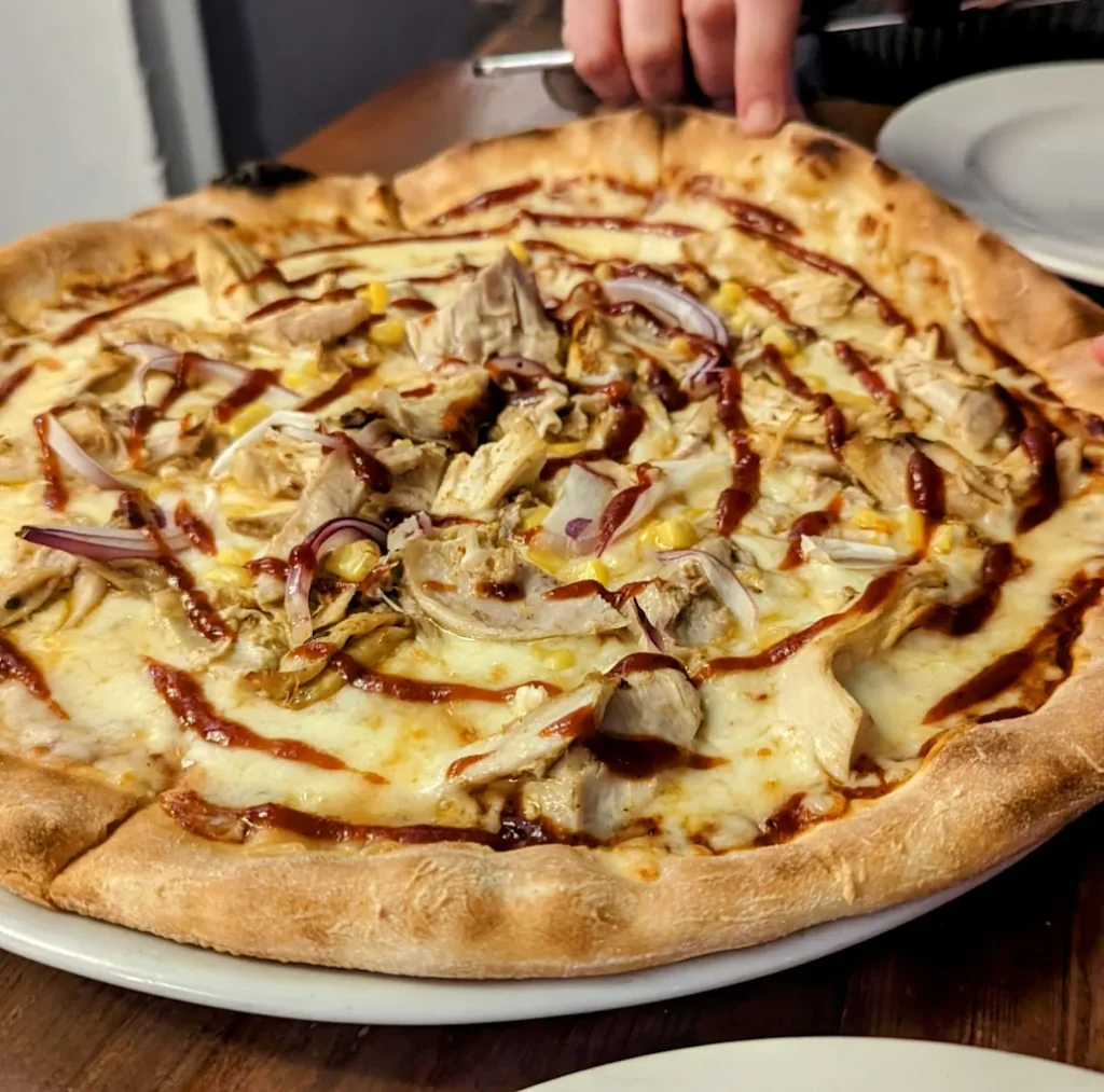 A large, golden-brown pizza with a crispy crust and melted cheese sits on a wooden table, surrounded by white plates. A hand holds a knife, ready to slice into the pizza. The scene is set in a cosy restaurant, with a blurred background emphasizing the pizza and table.