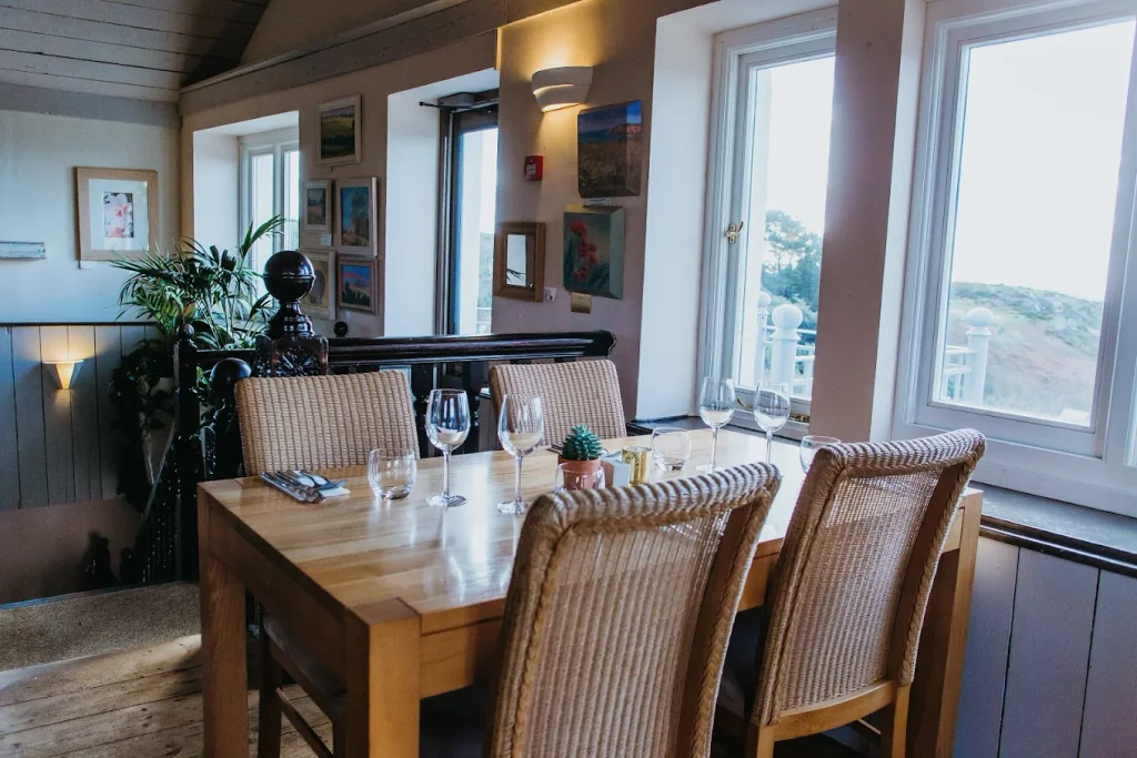The image depicts a cosy dining room with a wooden table set for four, surrounded by wicker chairs. The table is adorned with wine glasses, a salt shaker, and a green plant. The room features a large window with a view of a mountain, a white-framed painting, and a black globe. The walls are adorned with framed pictures and a lamp, creating a warm and inviting atmosphere.