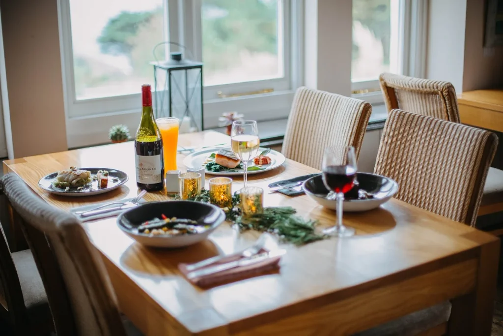 The image depicts a cosy dining scene with a large wooden table set for four, surrounded by chairs. The table is adorned with various dishes, glasses, and napkins, and is set against a window with a view of trees outside. The table is also decorated with a green wreath and a lantern, creating a warm and inviting atmosphere.