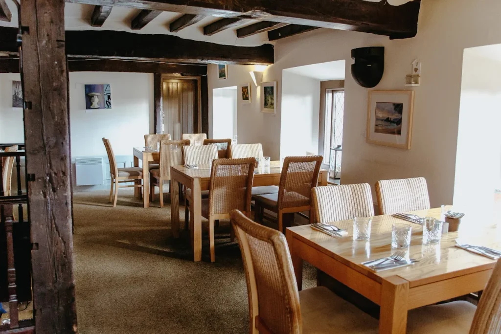 The image shows a cosy dining room with wooden tables, chairs, and a rug. The walls are adorned with paintings and a speaker, while a window allows natural light. The room is well-lit and has a warm, inviting atmosphere.