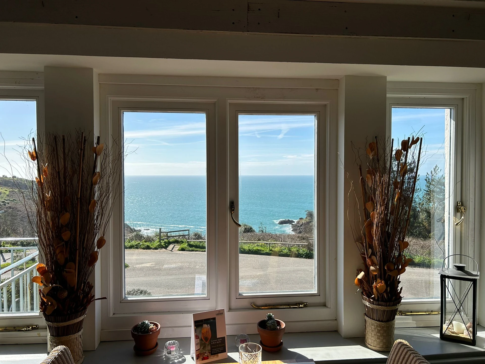 The image features a serene indoor scene with a large window offering a breathtaking ocean view. The window has white frames and two large, dry-bought plants with orange leaves. A white table with a vase of flowers and a candle is in front of the window, and a wooden shelf holds a photo and a candle. The room is well-lit and has a minimalist aesthetic, creating a tranquil atmosphere.