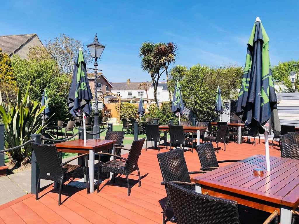 The image shows a serene outdoor patio area with wooden tables and chairs, green umbrellas, and a lamppost. The patio is surrounded by lush greenery and houses in the background. The scene is bathed in warm sunlight, creating a tranquil atmosphere.