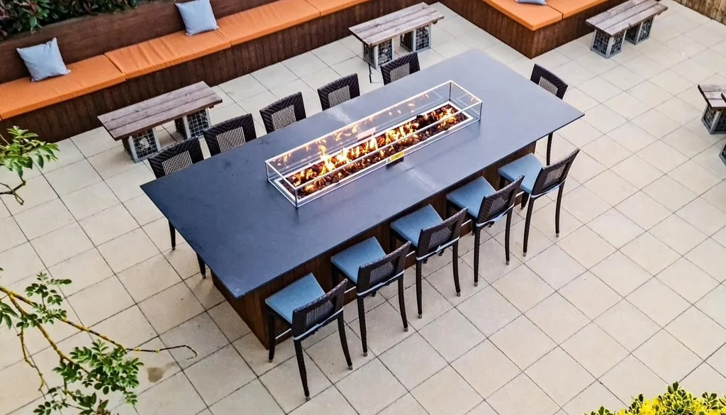 The image shows a tranquil outdoor patio area with a central black bar, six blue stools, and a wooden table with a fire pit. The patio is surrounded by wooden benches and a wooden bench with a blue cushion. The area is surrounded by a green lawn and trees, creating a serene and inviting atmosphere.