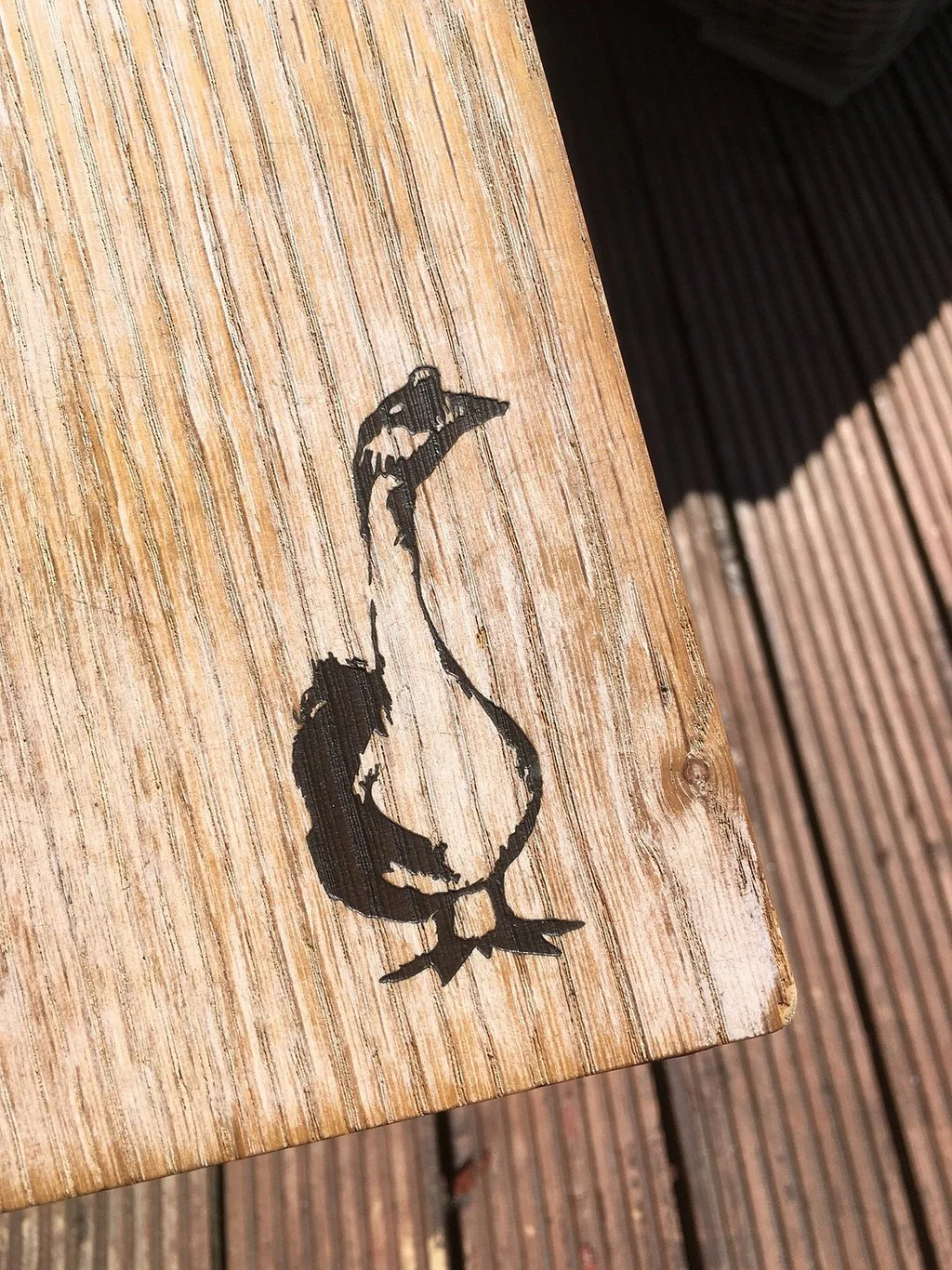A weathered wooden plank with a black duck silhouette on the right side is the main subject, set against a wooden deck with vertical slats. The image is taken from a low angle, emphasizing the plank's texture and the duck's silhouette.