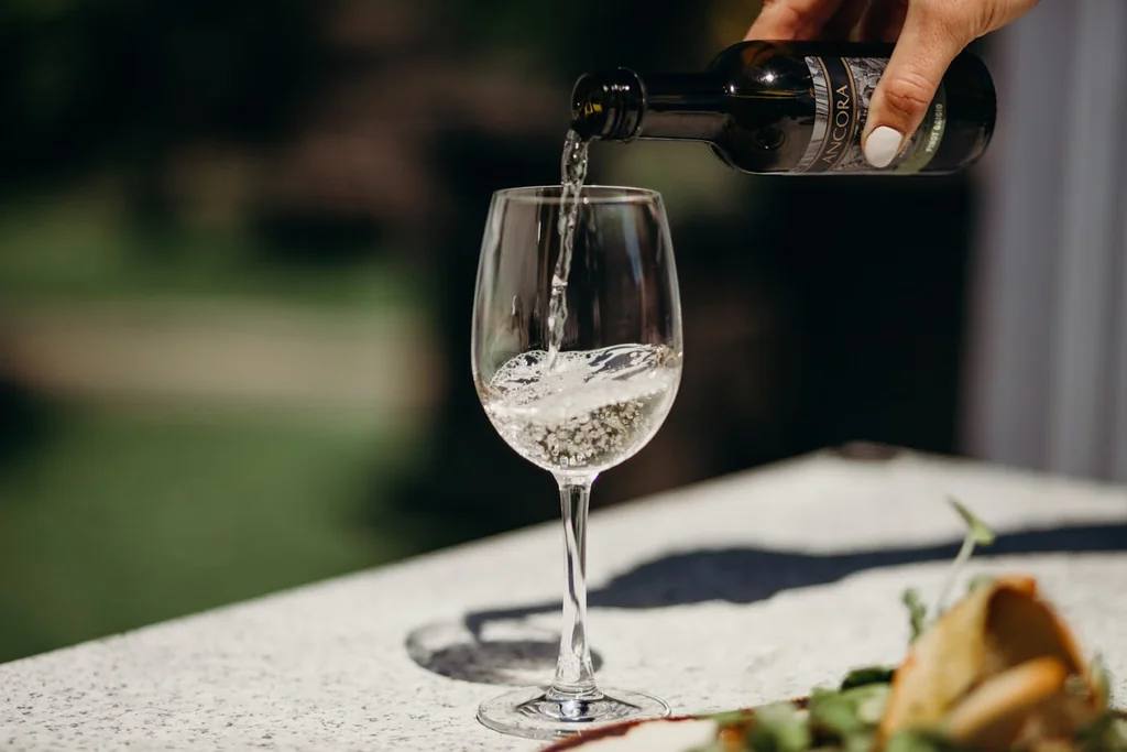 A hand pours white wine from a dark-colored bottle into a glass on a white tablecloth, with a green salad and bread nearby. The blurred background suggests an outdoor setting, possibly a garden or patio. The scene is serene and elegant, with the wine and salad as the main focus.