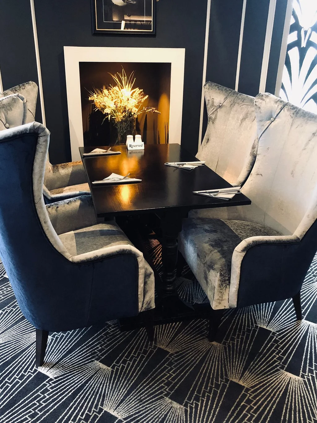 The image features a modern dining room with a black table, white and blue armchairs, and a white vase with yellow flowers. The table is surrounded by white napkins and a white candle. The room has a black and white geometric patterned floor and a black fireplace with a white frame. The objects are arranged harmoniously, creating a balanced and inviting atmosphere.