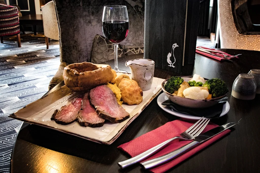 The image features a beautifully set dining table with a variety of food and utensils, set against a warm, inviting backdrop. The table is adorned with a wooden tray, a plate of meat, a bowl of potatoes, a glass of red wine, a white mug, a silver fork, a knife, and a red napkin. The table is set on a patterned rug, and the background includes a wooden chair and a fireplace. The scene exudes elegance and comfort, inviting the viewer to partake in the meal.