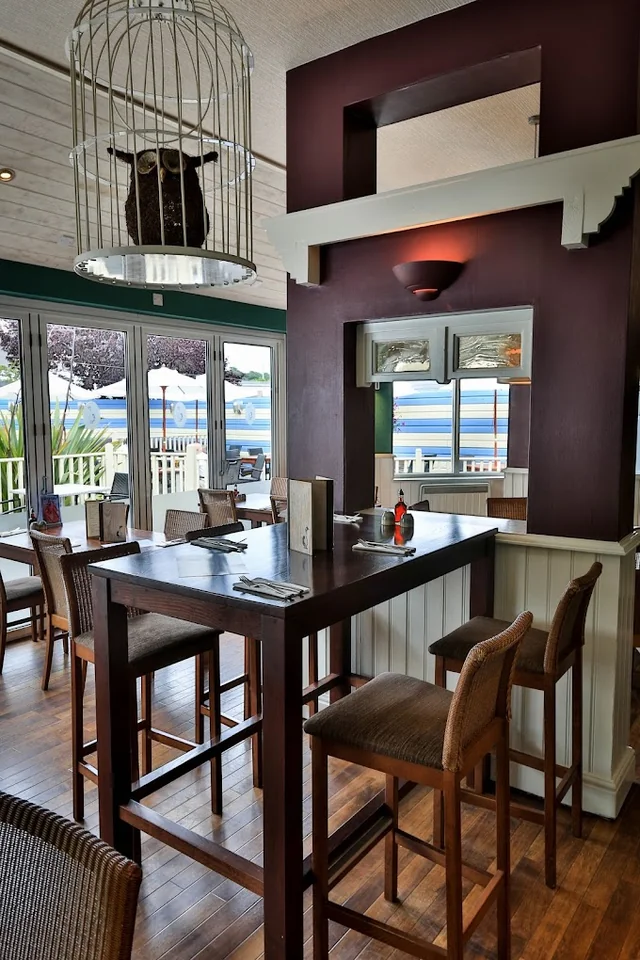 The image shows a modern restaurant with a wooden bar, four stools, and a white countertop. A red chandelier hangs from the ceiling, and a mirror reflects the view outside. The wooden floor and white walls create a warm and inviting atmosphere. The view from the window offers a serene view of the ocean, and the restaurant is well-lit and organized.