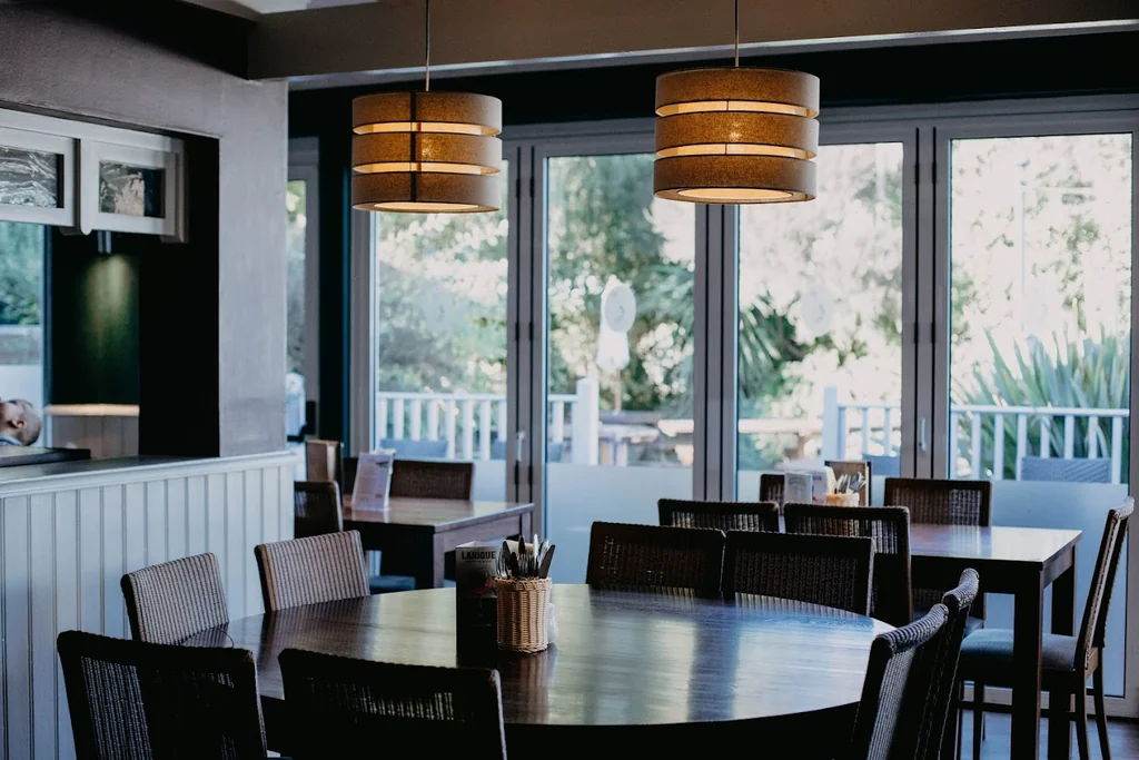 The image shows a modern, minimalist restaurant with a large wooden table, six chairs, and a vase with a salt and pepper shaker. The room has a neutral color palette, with a white wall and black ceiling. Large windows offer a view of trees outside, and pendant lights hang from the ceiling. The restaurant is well-lit and has a welcoming atmosphere.