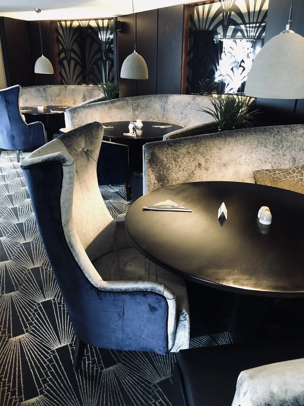 The image features a modern, sophisticated restaurant with a black and white geometric patterned floor, a black and silver bar table with a black top and silver legs, and blue velvet armchairs. The tables are adorned with white candles and silverware, and the walls are adorned with black and white geometric patterns. The restaurant is illuminated by white pendant lights, creating a warm and inviting atmosphere.