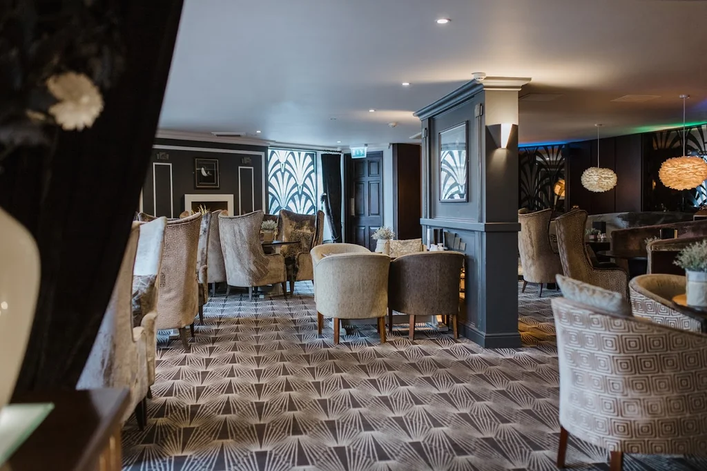 The image features a luxurious lounge area with a black and white patterned rug, beige chairs, and tables, a bar with a wooden top and white walls, and a large window with white curtains. The room is illuminated by hanging lights and a large chandelier, creating a sophisticated and inviting atmosphere.