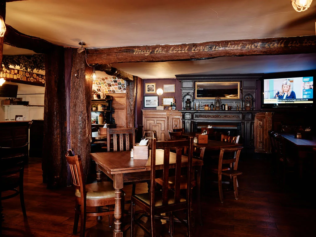 The image depicts a cozy, rustic restaurant with a warm, inviting atmosphere. The wooden floor and walls create a rustic charm, while the wooden furniture and chairs add to the cozy ambiance. A large television is mounted on the wall, displaying a news program. The room features a fireplace, a wooden bar, and a wooden cabinet with a mirror. The lighting is warm and inviting, with hanging lights and a chandelier. The scene is filled with details, creating a sense of comfort and relaxation.