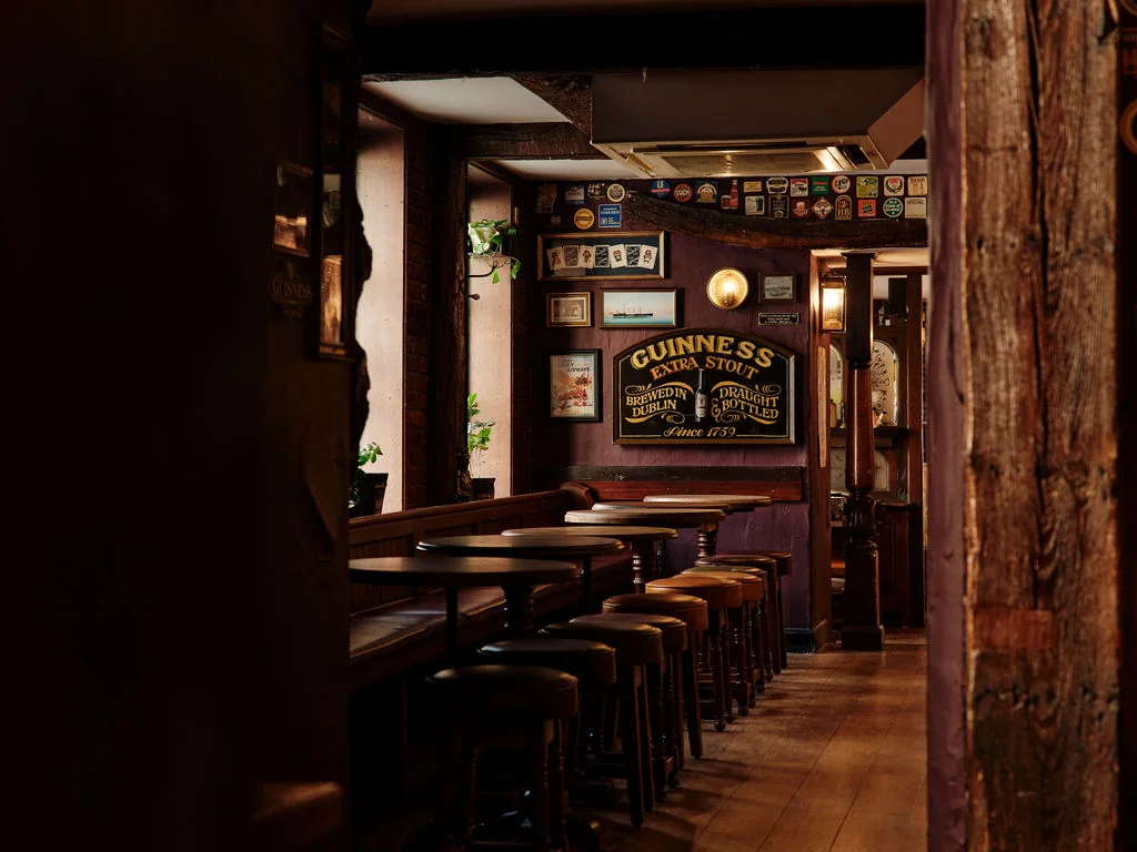 The image shows a cozy, dimly lit bar with wooden tables and stools, a wooden bar, and a wooden door leading to the outside. The walls are adorned with various items, including a clock, a sign, and a plant. The atmosphere is warm and inviting, with a sense of history and nostalgia.
