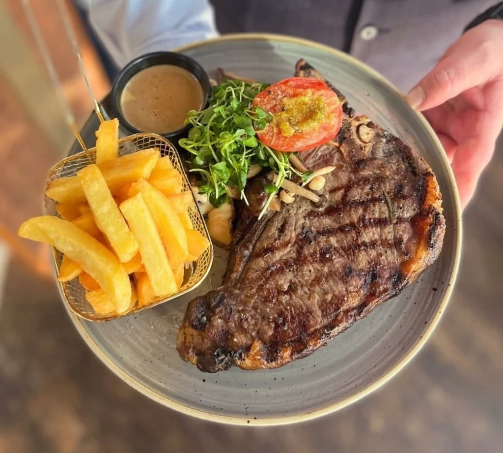 A person holds a gray plate with a steak, fries, and salad, surrounded by a wooden table and a black shirt. The steak is grilled to perfection, with a grilled tomato and a slice of red onion. The fries are golden and crispy, and the salad is fresh and vibrant. The scene is visually appealing and suggests a satisfying meal.