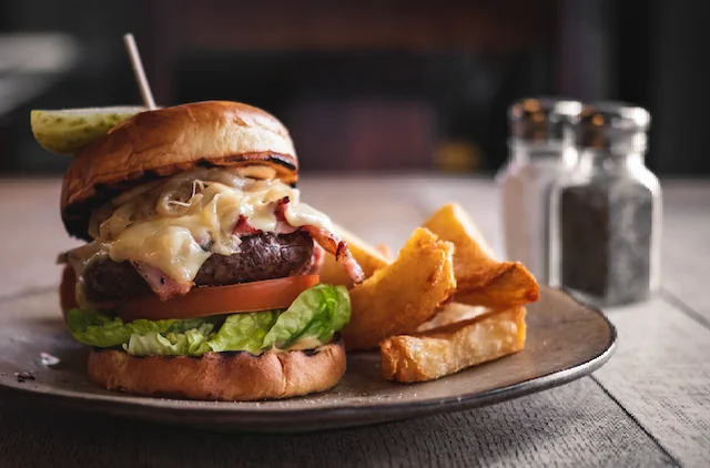 A large, golden-brown burger with a bun, lettuce, tomato, cheese, and pickle is the centerpiece of a meal on a round metal plate. The burger is accompanied by golden-brown potato chips. The plate sits on a wooden table, with a salt shaker and pepper shaker nearby. The background is blurred, focusing on the meal.