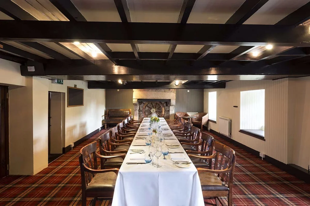 The image shows a spacious, well-lit dining room with a long, white-clothed table set for eight, surrounded by wooden chairs and a fireplace. The room features a red and black checkered carpet, white walls, and a high ceiling with dark wood beams. The arrangement of objects and the precise location of each object contribute to a harmonious and inviting atmosphere.