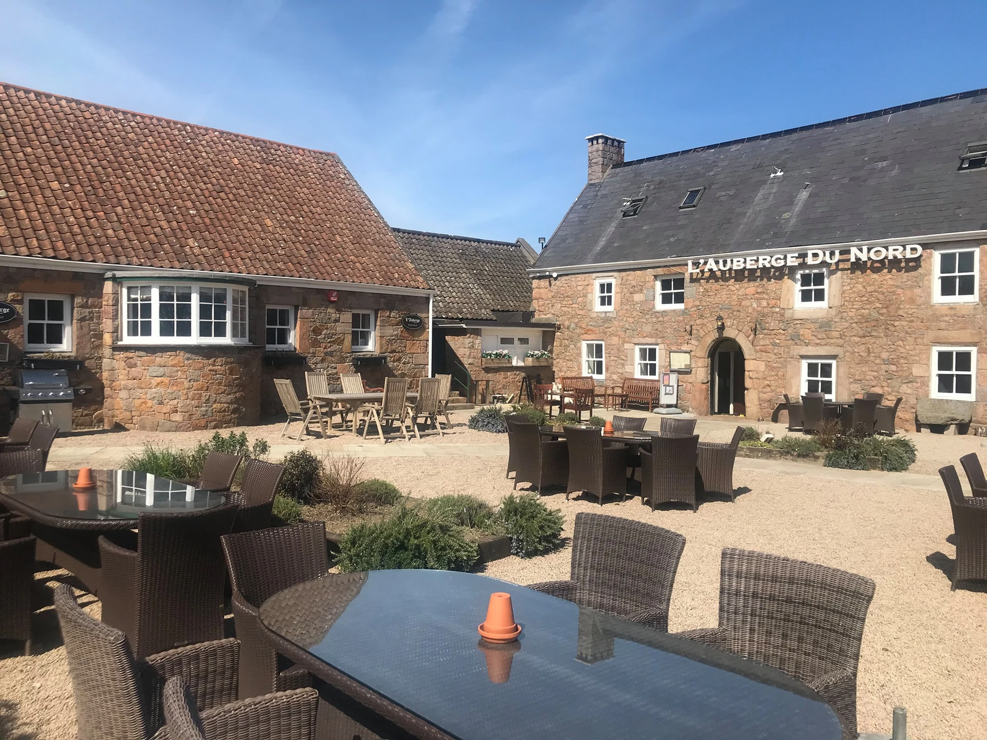 The image shows the "Huis de Brouwer" pub in the Netherlands, with a brick building featuring a red tiled roof and a sign. The pub is located on a gravel courtyard, with tables and chairs arranged for patrons. The scene is set against a clear blue sky, creating a serene atmosphere.