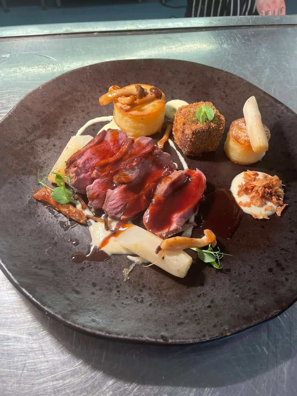 A black plate with a variety of food items is on a metal countertop, featuring a piece of dark meat, a fried egg, a fried onion, a small meat ball, and a small meat ball with a sauce. The plate is set against a blurred background, emphasizing the food's presentation.