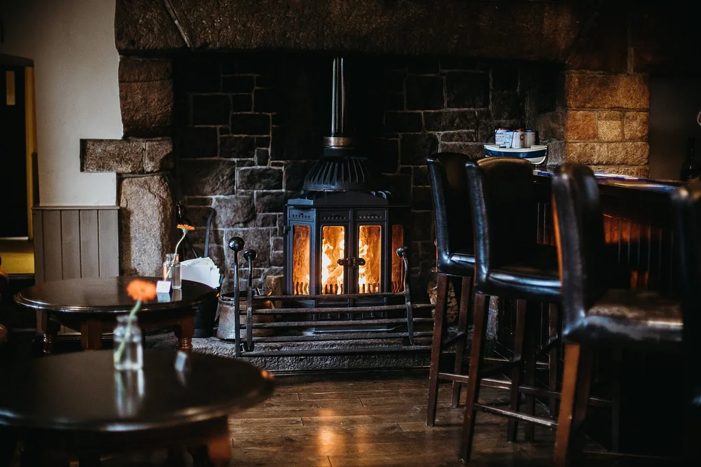 A cozy bar area features a dark stone fireplace with a lit fire, casting a warm glow. A wooden table with a vase of flowers and a wooden stool are in front of the fireplace. Bar stools are to the right, and a wooden bar is on the left. The room has a warm, inviting atmosphere with a wooden floor and a stone wall.